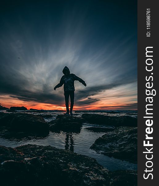 Person Wearing Hoodie Standing on Rock Surrounded by Body of Water