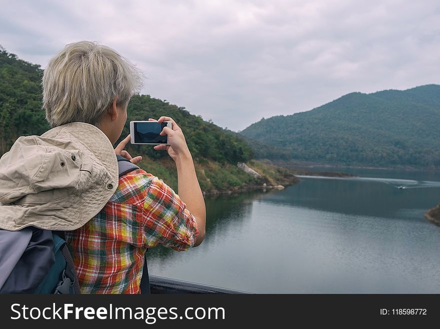 Person Holding Smartphone While Taking Photo of Calm Water Near Mountain at Daytime