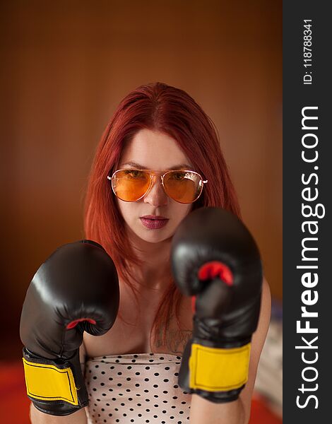 Pretty young woman with red hair, sunglasses and boxing gloves looks upset, feminism, self defense concept