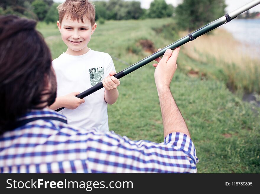 A picture of happy son standing in front of his dad and holding long fish-rod. Guy is holding it a bit. They are spending time together