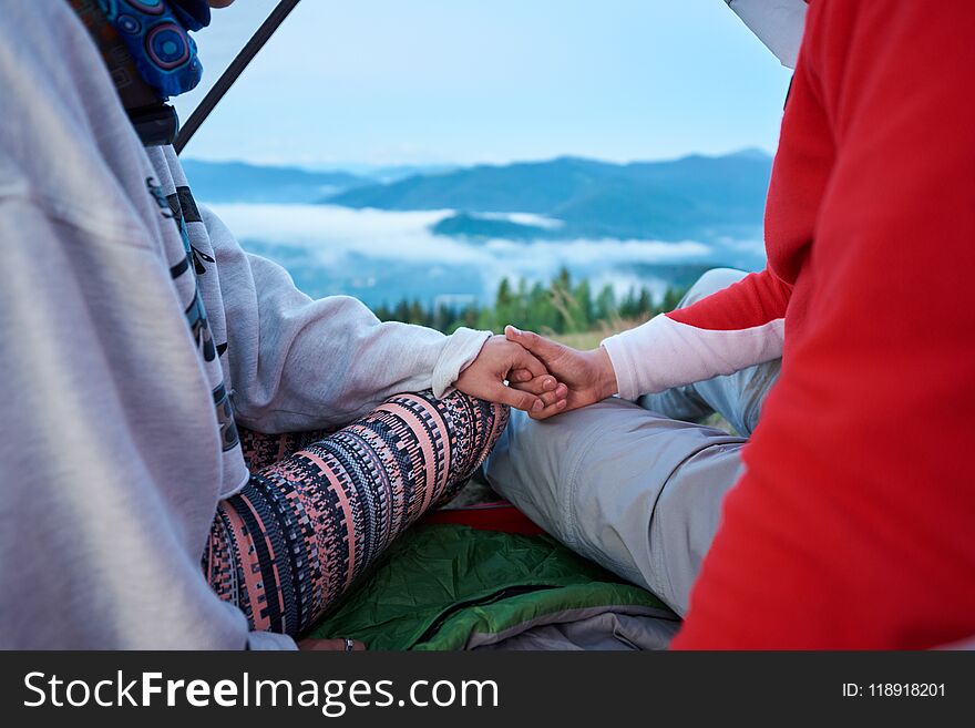 Close-up of guy and girl holding hands sitting in tent on blurred background mountains