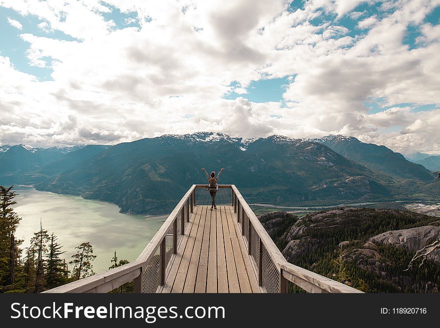 Person Standing With Hands on Air on Brown Wooden Dock With Overlooking View of Lake Under White Clouds and Blue Sky