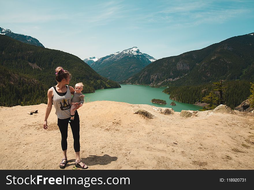 Woman Wearing Grey Tank Top Carrying Baby in Distant of Lake Between Mountains