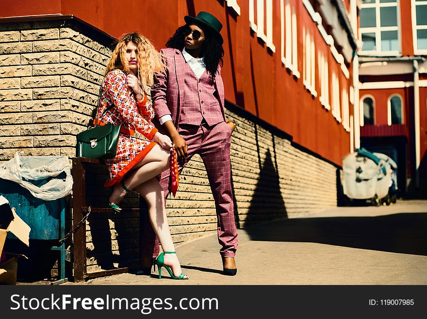 Shallow Focus Photography Of Woman In Orange Dress Leaning On Brown Wall Beside Man In Purple Suit Jacket