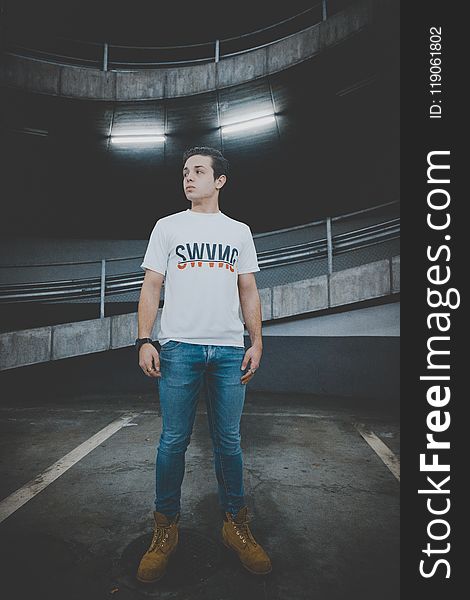 Man Wearing White T-shirt and Blue Jeans Standing on Black Concrete Floor