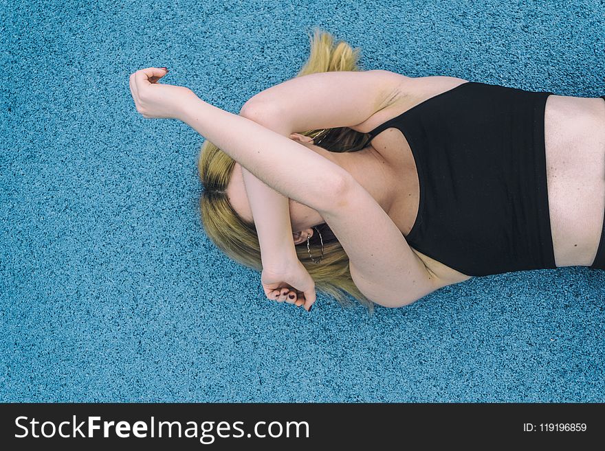 Woman Wearing Black Sports Brassiered Lying on Teal Surface While Covering Her Face With Her Arms