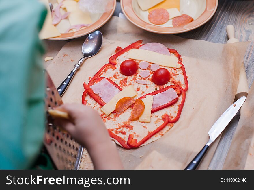 The child decorates the funny pizza - pepper, cheese, sausage, sauce spread out on the dough.