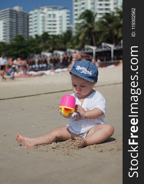 A Child In Sun-protective Clothes And A Headdress Plays On A Sandy Beach On A Clear Hot Day, Vertical Photo.