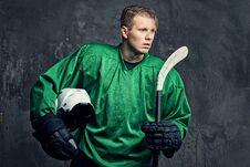 Tired Professional Hockey Player In A Green Sportswear Holds A Hockey Stick And Protective Helmet On A Gray Background. Royalty Free Stock Photo