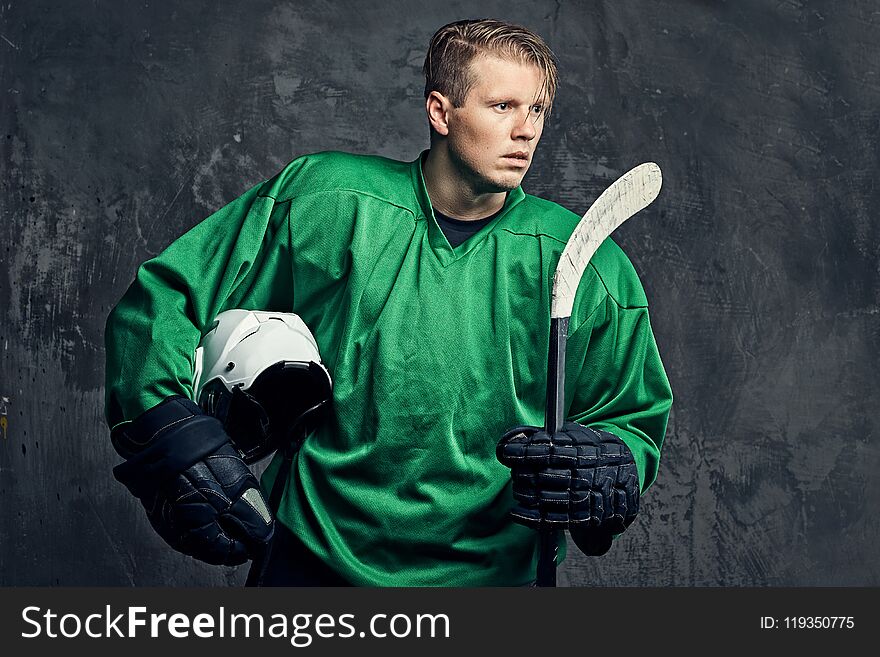 Tired professional hockey player in a green sportswear holds a hockey stick and protective helmet on a gray background.