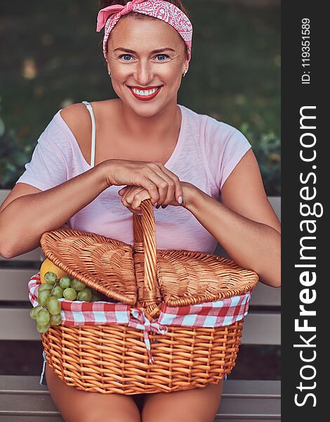 Happy beautiful redhead female wearing casual clothes holds a picnic basket while sitting on a bench in a park.