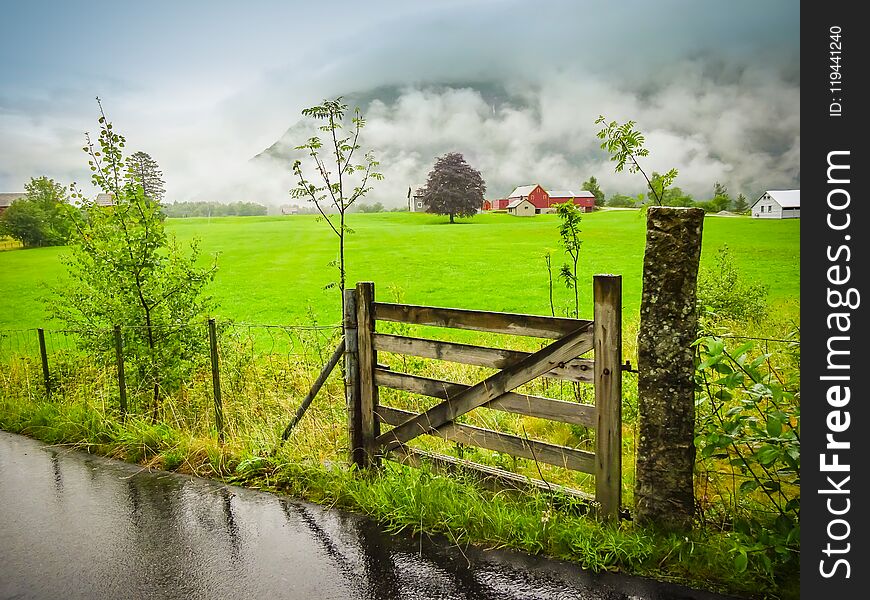 Countryside with farm and road on a rainy summer day. Wooden gate and fence with beautiful green grass field. Norway, Scandinavia. Countryside with farm and road on a rainy summer day. Wooden gate and fence with beautiful green grass field. Norway, Scandinavia.