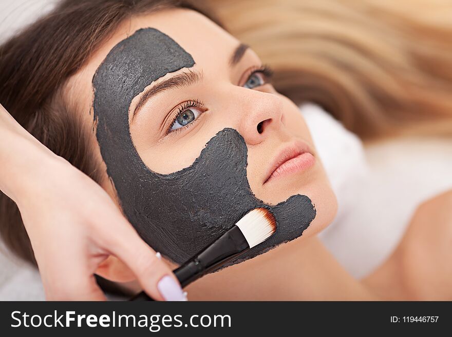 People, beauty, spa, cosmetology and skincare concept - close up of beautiful young woman lying with closed eyes and cosmetologist applying facial mask by brush in spa.