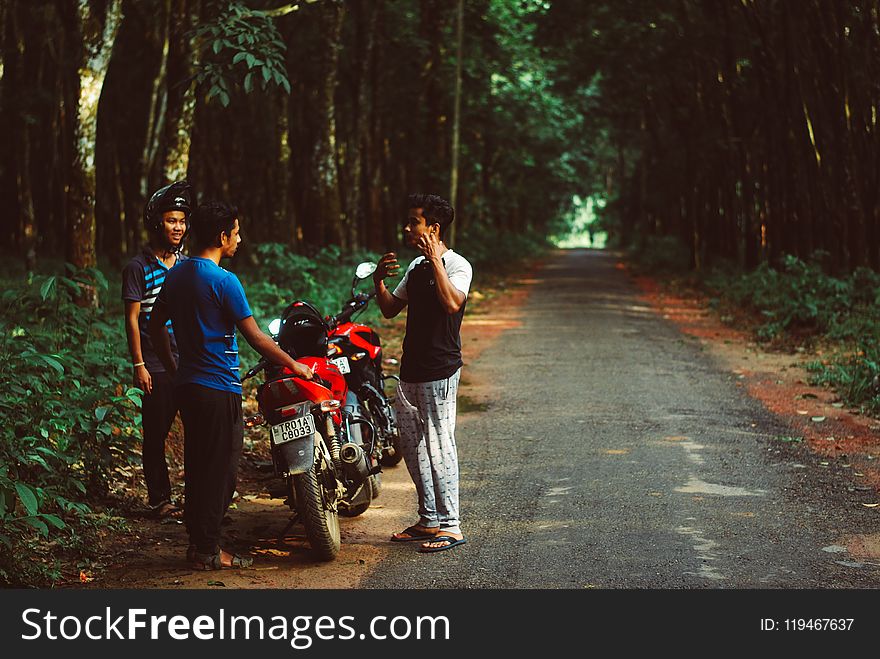 Three Men Standing Beside Red Motorcycle Surrounded With Green Trees