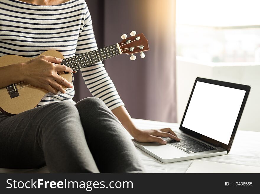 Happy young asian woman playing ukulele sitting on bed in bedroom.