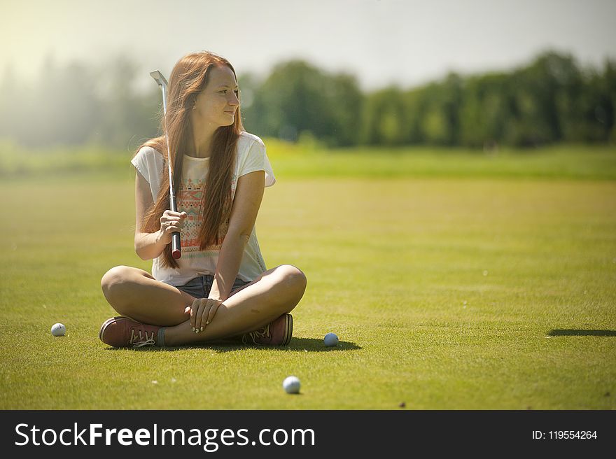 Woman In White Scoop-neck Shirt And Blue Shorts Holding A Golf Club Sitting On Golf Field