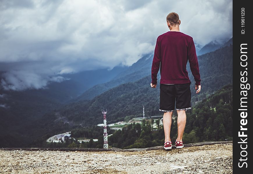 Man Wearing Maroon Sweater and Black Shorts Standing in Front of Mountain