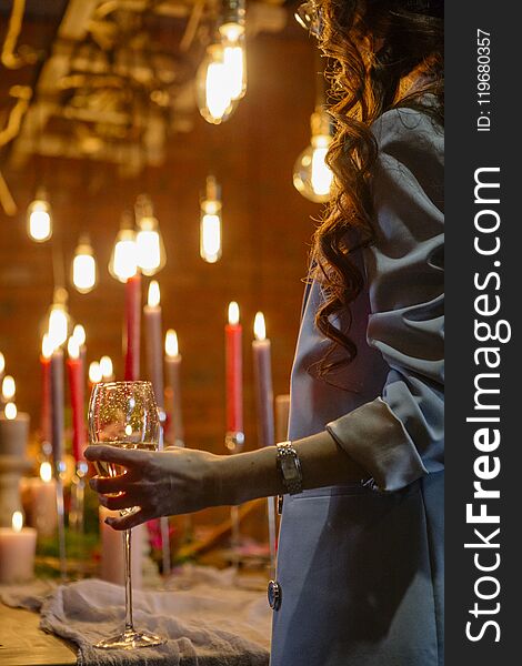 Woman standing neat wedding table, celebrate with a glass of wine in her hand. No face. Decor, candles and edison lamps on a background
