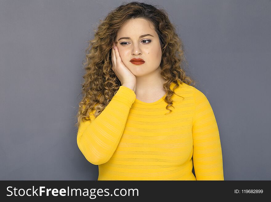 Cute brunette plus size woman with curly hair in yellow sweater and jeans standing on a neutral grey background. She has a thooth pain, holding her hand on her cheek