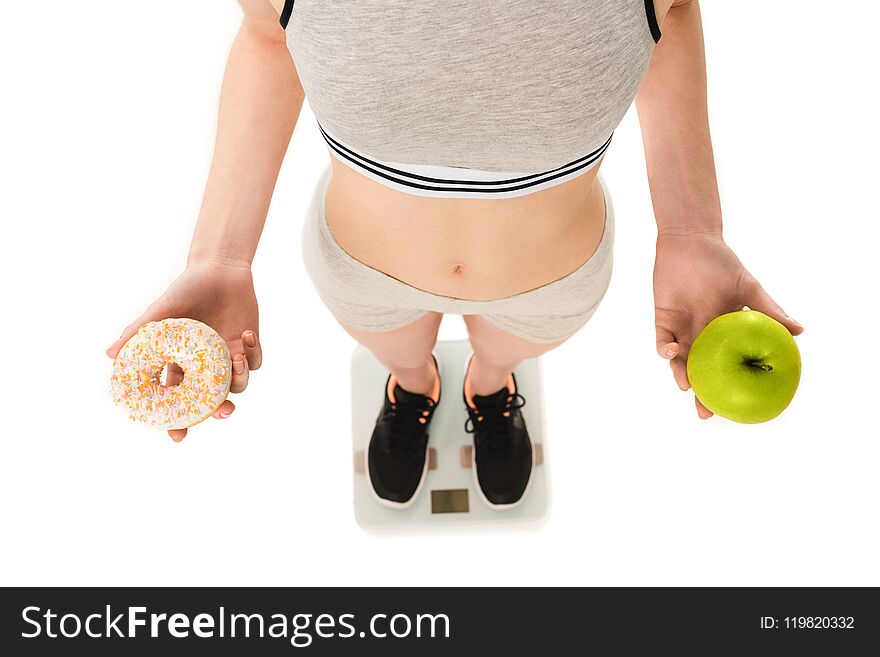 cropped shot of slim woman with apple and doughnut standing on scales isolated on white