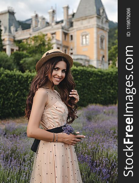 Fashion photo of beautiful woman with dark hair in elegant dress and hat posing in flowering lavender field near antic castle