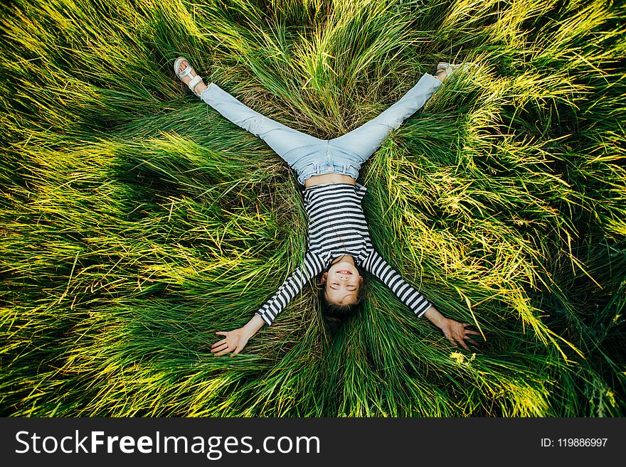 Little girl in a striped T-shirt lies on a big green glade and looks at the camera smiling. Top view. Little girl in a striped T-shirt lies on a big green glade and looks at the camera smiling. Top view.