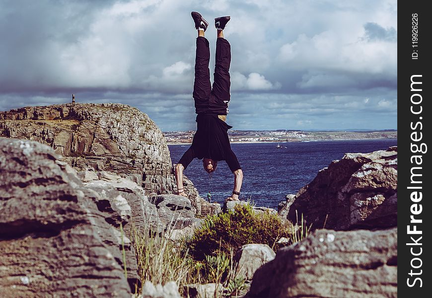 Man Wearing Black Dress Shirt and Burgundy Pants Hand Standing on Rock Formations Across Sea