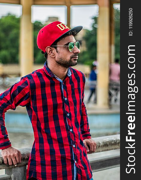 Selective Focus Photography of Man in Red and Black Gingham Sports Shirt Leaning on Grey Concrete Fence