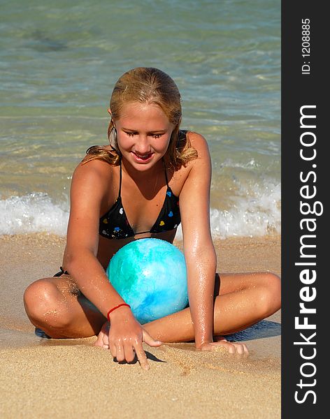 A teenager girl with a blue ball is sitting on a sandy beach near the water. A teenager girl with a blue ball is sitting on a sandy beach near the water