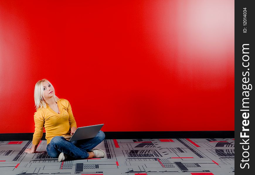 Young blond woman sitting on the floor with the computer on her knees, red wall background. Young blond woman sitting on the floor with the computer on her knees, red wall background