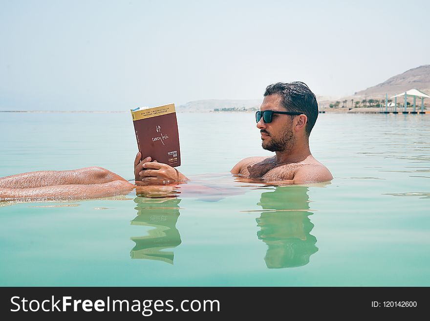 Man Wearing Sunglasses Reading Book on Body of Water