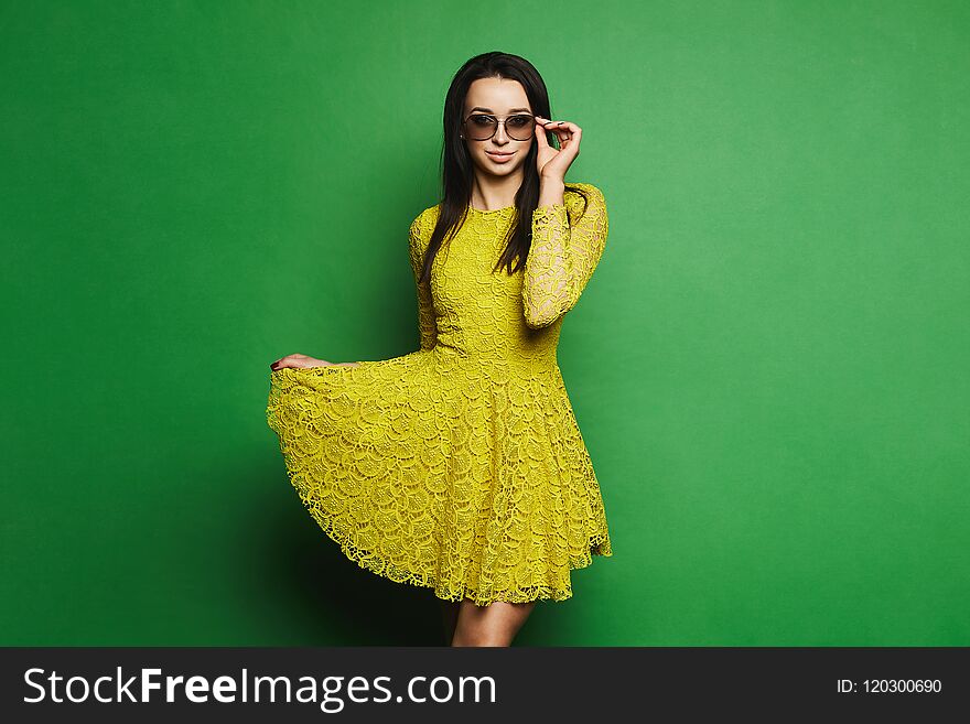 Beautiful, and fashionable brunette model girl in short yellow dress and sunglasses pulls up the hem of her dress and posing