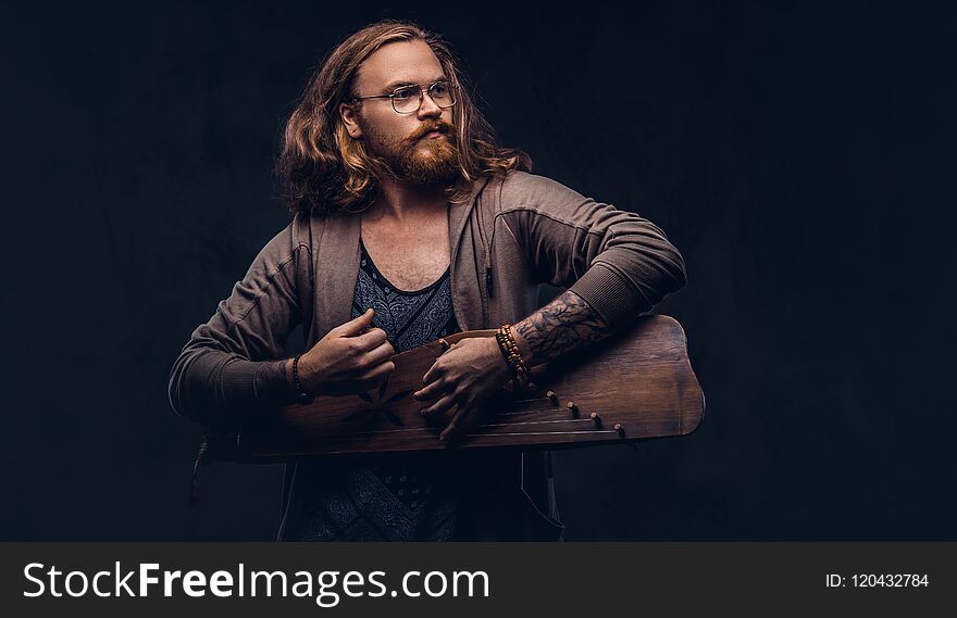 Redhead hipster male with long luxuriant hair and full beard dressed in casual clothes playing on a Russian traditional musical instrument - gusli. Isolated on the dark background.