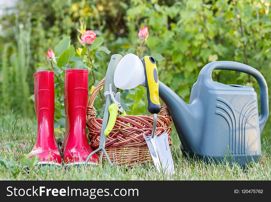 Red garden rubber boots, small rake, pruner, wicker basket, trowel and plastic watering can on green grass with blurred green background. Tools for gardening