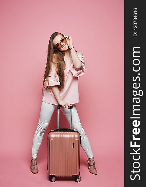 Beautiful blonde model girl in fashionable blouse with naked shoulders and in stylish sunglasses stands with suitcase and posing at pink background in studio.