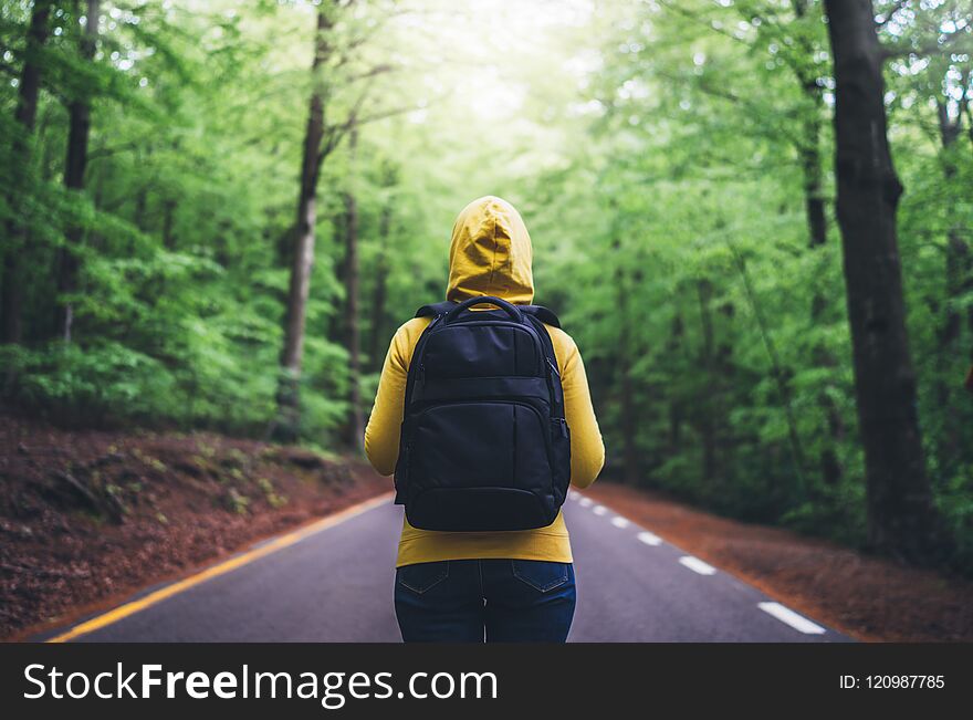 Tourist traveler with backpack standing into road at summer green forest, view back girl hiker in yellow hoody looking and enjoyin