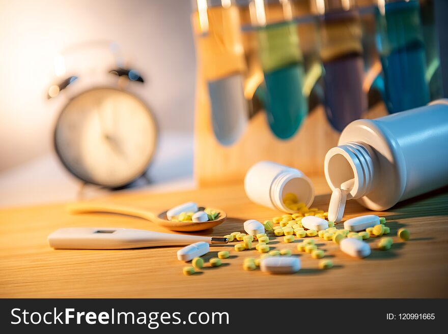 Pills spilling out of pill bottles with digital thermometer and pharmaceutical spoon on wooden board. Table clock and test tube in the background. Prescription medicine and medical treatment concepts. Pills spilling out of pill bottles with digital thermometer and pharmaceutical spoon on wooden board. Table clock and test tube in the background. Prescription medicine and medical treatment concepts