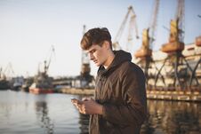 Young Man With Brown Hair Standing And Thoughtfully Looking In His Mobile Phone. Cool Boy In Down Jacket Standing With Stock Photo