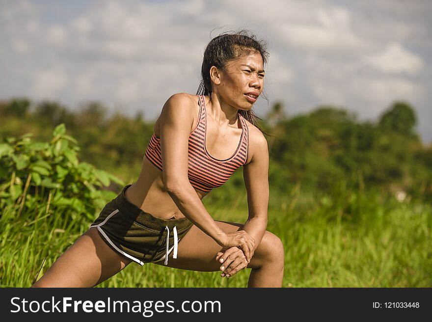 Fit and sporty middle aged runner Asian woman stretching leg and body after running workout on green field beautiful background in