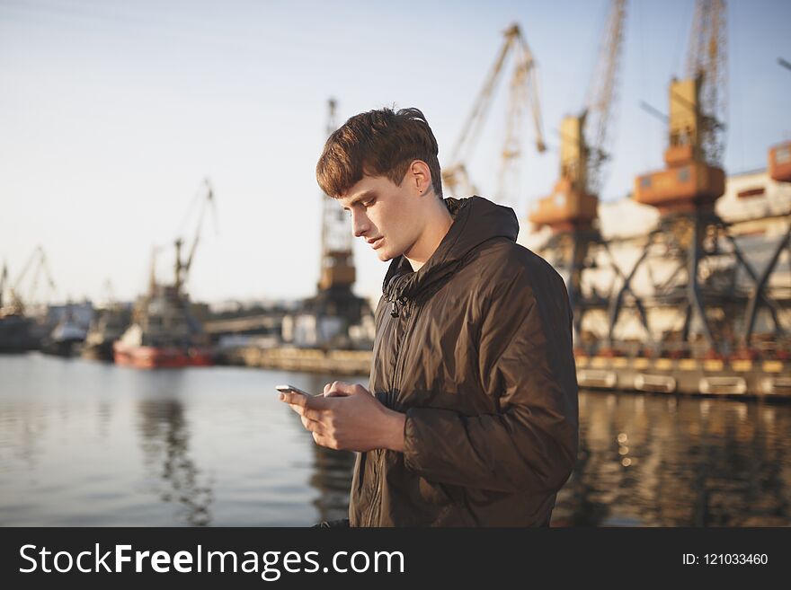 Portrait of young man with brown hair standing and thoughtfully looking in his mobile phone. Cool boy in down jacket standing with cellphone in hands with sea port on background