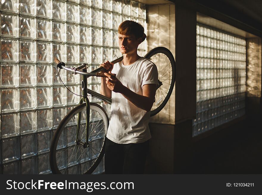 Portrait of thoughtful boy standing and holding bicycle that leaning on his shoulder while dreamily looking aside. Young man in white t-shirt standing with classic bicycle