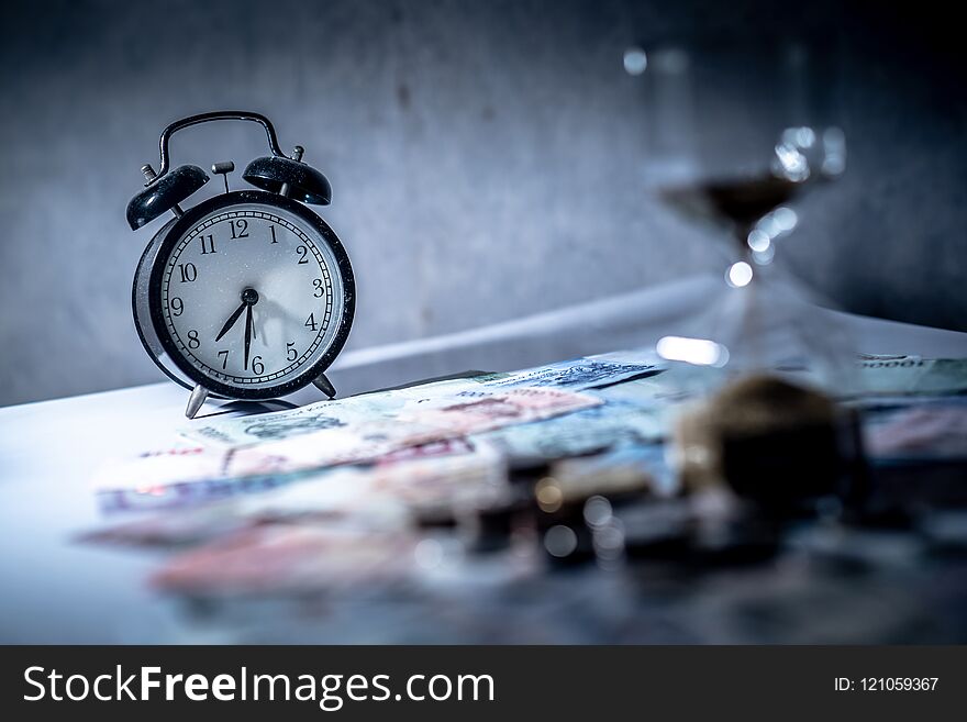 Reflection of clock and blurred hourglass on glowing table with banknotes and coins of international currency. Time investment and passing time. Urgency countdown timer for business deadline concept. Reflection of clock and blurred hourglass on glowing table with banknotes and coins of international currency. Time investment and passing time. Urgency countdown timer for business deadline concept