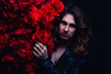 Brutal Man In A Leather Jacket On A Naked Body With Long Hair Stands Near The Decor With Red Flowers Flowers. Strength Royalty Free Stock Image