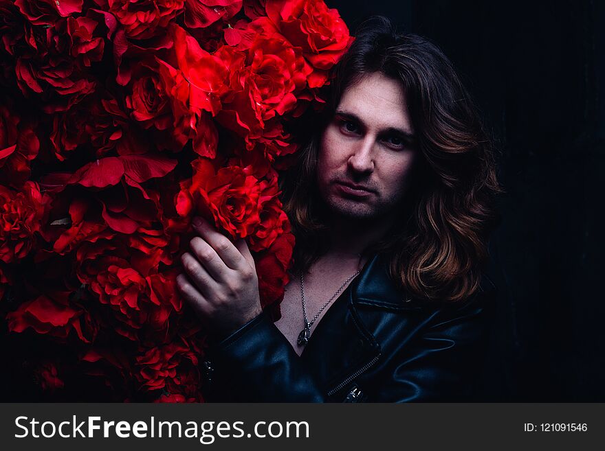 Brutal man in a leather jacket on a naked body with long hair stands near the decor with red flowers flowers. strength
