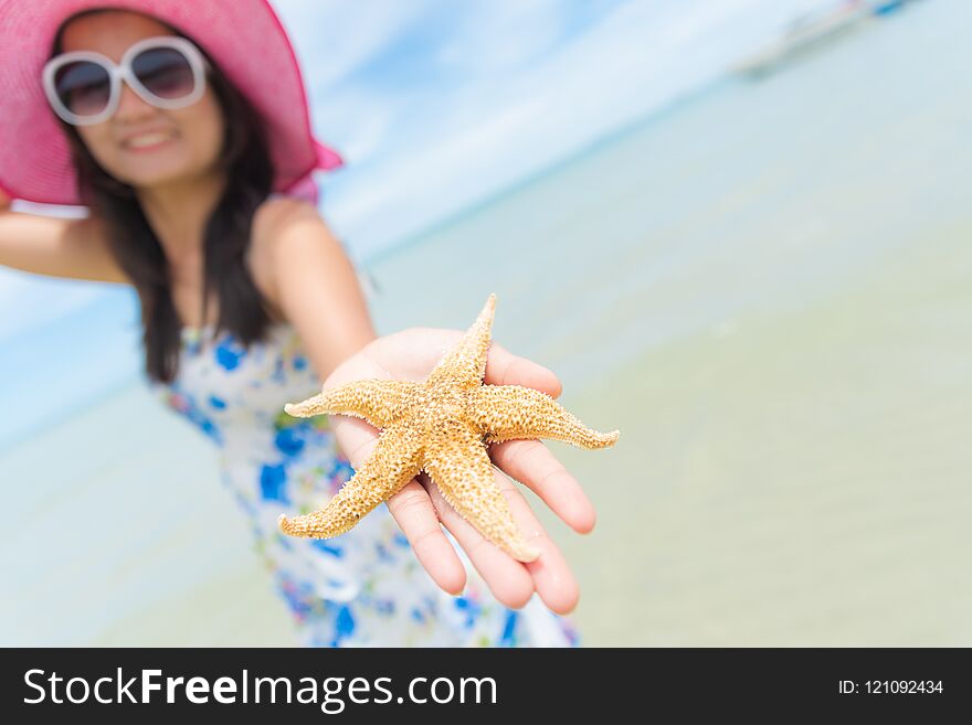 Beautiful woman wearing hat beach and sunglasses and holding starfish on hands over sandy beach, green sea and blue sky background for summer holiday and vacation concept.