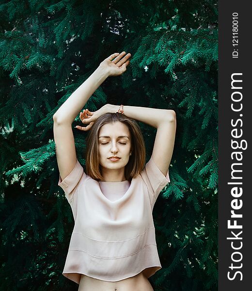 Lifestyle portrait of young white woman with short hair standing in front of pine tree with her eyes closed and hands lifted, concept of natural beauty or closeness-to-nature