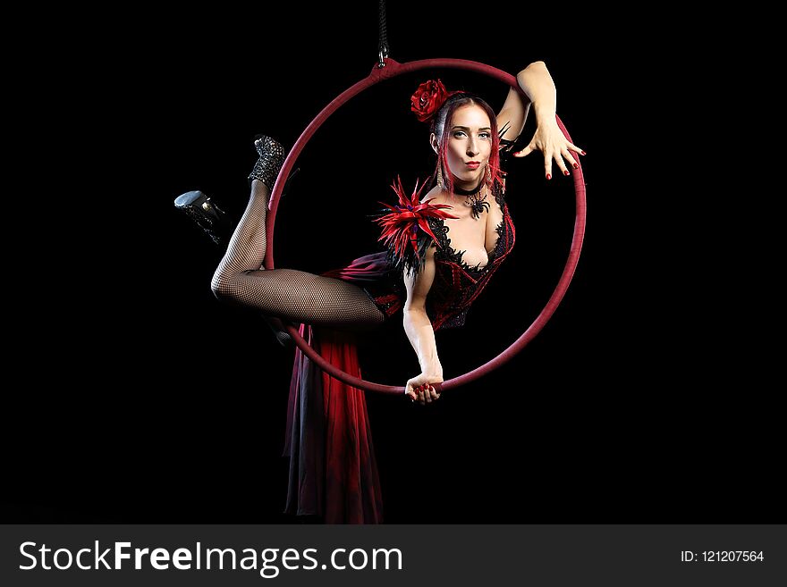 A adul girl in red evening dress performs the acrobatic elements in the air ring on a black background.