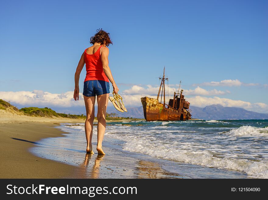 Travel freedom. Mature tourist woman walking on beach enjoying summer vacation. An old abandoned shipwreck, wrecked boat in the background. Travel freedom. Mature tourist woman walking on beach enjoying summer vacation. An old abandoned shipwreck, wrecked boat in the background