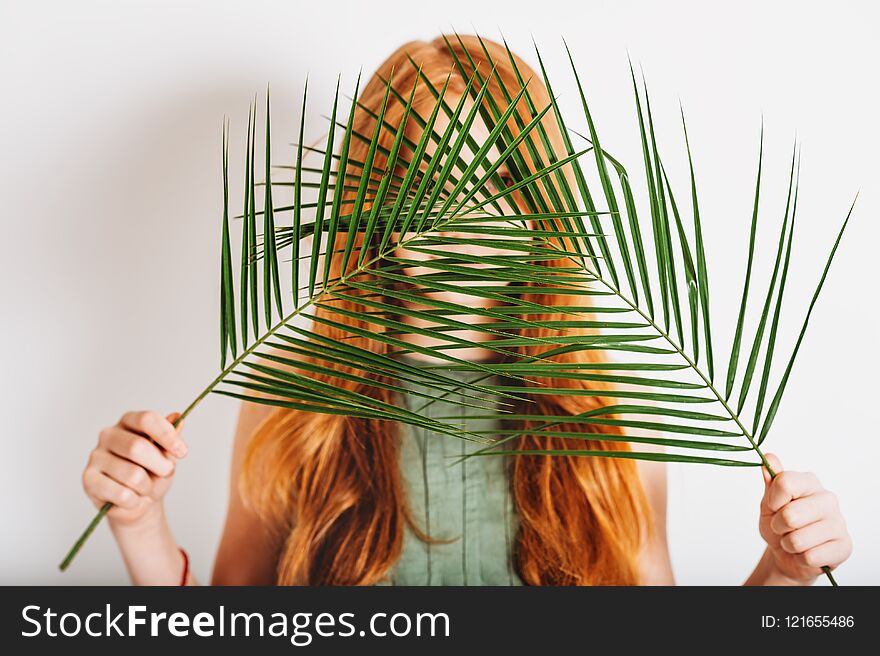 Studio shot of adorable red-haired preteen kid girl, wearing khaki dress, hiding behind exotic green palm leaves. Studio shot of adorable red-haired preteen kid girl, wearing khaki dress, hiding behind exotic green palm leaves