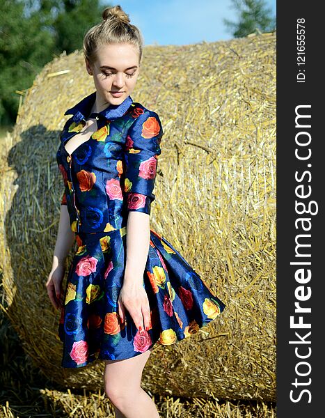 Female model, outdoor photo session, farmland in summer. Girl in colorful dress stands in front of roll of hay. Female model, outdoor photo session, farmland in summer. Girl in colorful dress stands in front of roll of hay.